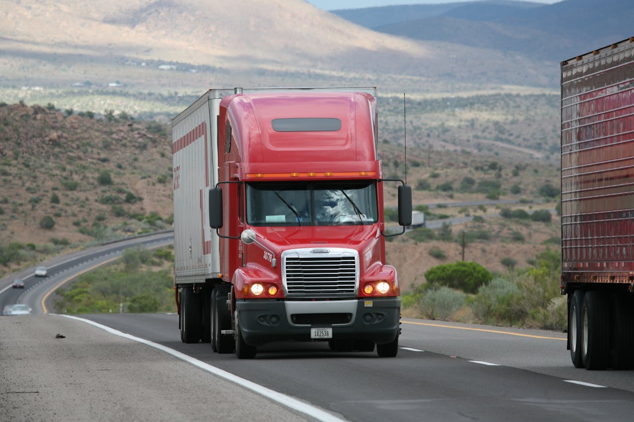 Red semi-truck driving through scenic highway in a mountainous landscape.